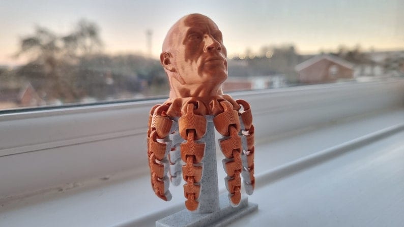 Op de afbeelding: Een 3D-geprint buste van een mannenhoofd met een koperen afwerking. De buste is gemonteerd op een basis met meerdere scharnierende armen.
