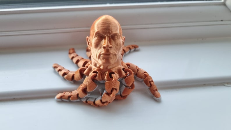 Op de afbeelding: Een 3D-geprint beeldje van een mensenhoofd met een serieuze uitdrukking, bevestigd aan het lichaam van een octopus. Het beeldje is gemaakt van koperkleurig materiaal.