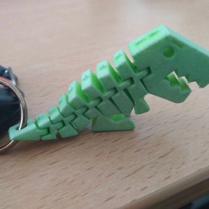 Puede incluir: Un llavero de pl&aacute;stico verde en forma de esqueleto de dinosaurio. El dinosaurio est&aacute; en posici&oacute;n de caminar con la cabeza girada hacia un lado. El llavero est&aacute; unido a un anillo plateado.