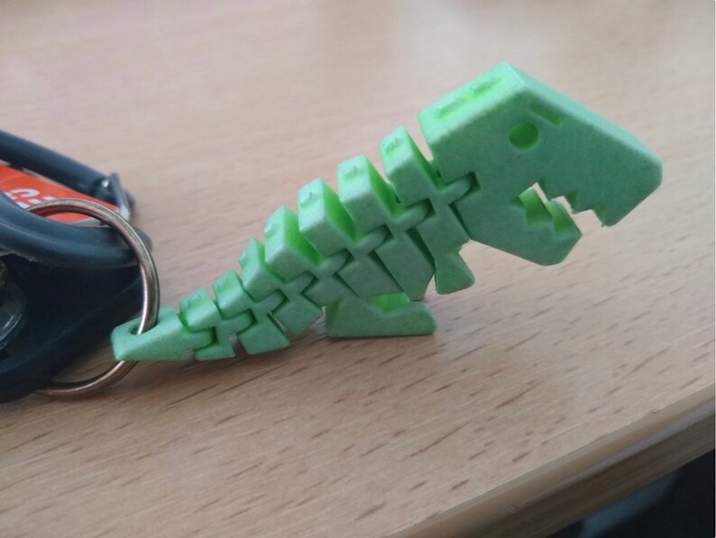 Puede incluir: Un llavero de pl&aacute;stico verde en forma de esqueleto de dinosaurio. El dinosaurio est&aacute; en posici&oacute;n de caminar con la cabeza girada hacia un lado. El llavero est&aacute; unido a un anillo plateado.