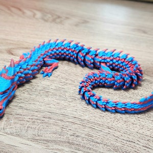 Puede incluir: Una figurita de drag&oacute;n impresa en 3D de color azul y rojo. El drag&oacute;n est&aacute; enroscado en una posici&oacute;n de descanso con la cabeza apoyada en la cola.