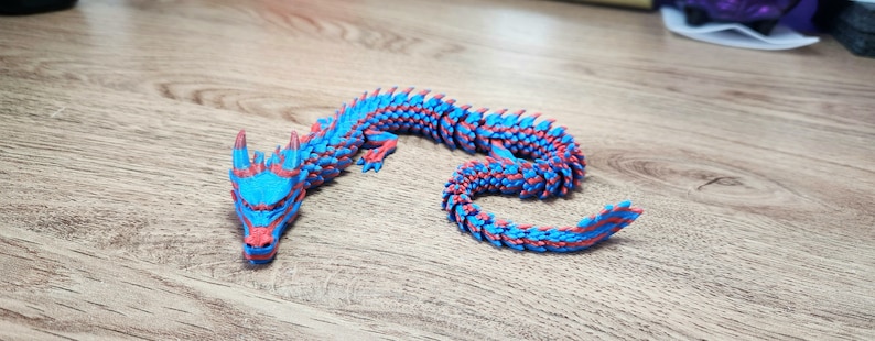 Puede incluir: Una figurita de drag&oacute;n impresa en 3D de color azul y rojo. El drag&oacute;n est&aacute; enroscado en una posici&oacute;n de descanso con la cabeza apoyada en la cola.