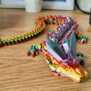 May include: A multi-colored origami dragon made of paper. The dragon is in a curled position with its head resting on the table. The dragon is made of many small, folded pieces of paper that are arranged in a pattern to create the dragon's scales. The dragon is a vibrant rainbow of colors, including red, orange, yellow, green, blue, indigo, and violet.