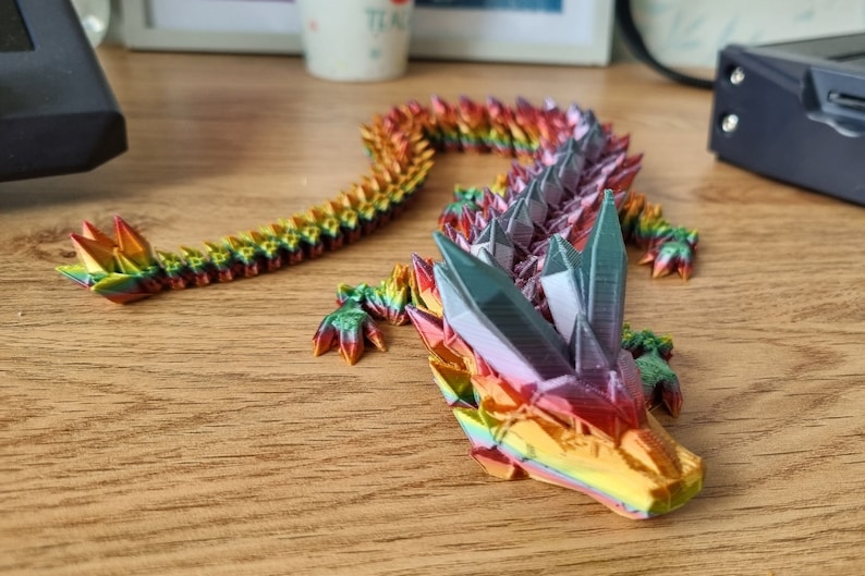 May include: A multi-colored origami dragon made of paper. The dragon is in a curled position with its head resting on the table. The dragon is made of many small, folded pieces of paper that are arranged in a pattern to create the dragon's scales. The dragon is a vibrant rainbow of colors, including red, orange, yellow, green, blue, indigo, and violet.