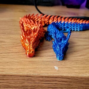 May include: Two small plastic dragons, one orange and one blue, are posed on a wooden surface. The dragons are detailed with scales and have a textured finish.