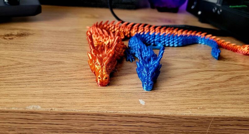 May include: Two small plastic dragons, one orange and one blue, are posed on a wooden surface. The dragons are detailed with scales and have a textured finish.