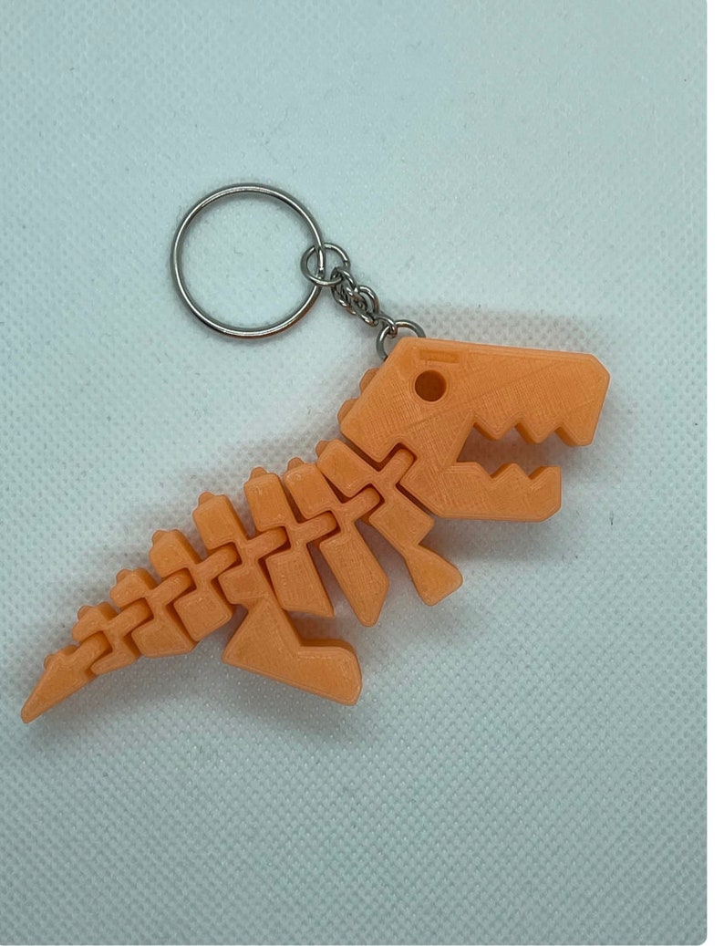 Puede incluir: Un llavero de pl&aacute;stico de esqueleto de dinosaurio de color melocot&oacute;n con un anillo y una cadena de metal plateado.