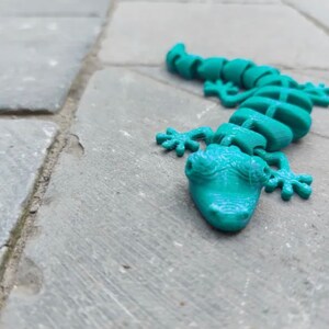 Peut inclure: Un jouet en forme de lézard articulé de couleur turquoise est posé sur une surface en pierre grise. Le lézard présente un design détaillé avec des segments visibles et de petits pieds. Le jouet mesure environ 20 cm de long.