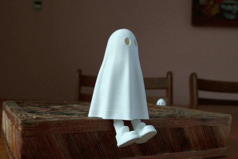 May include: A white ghost figurine with a sheet-like body and two small holes for eyes. The ghost is sitting on a wooden surface with its legs crossed.