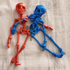May include: Two articulated skeleton figures, one red and one blue, are lying on a white fabric surface with a floral pattern. The skeletons are posed in a relaxed, almost playful manner.