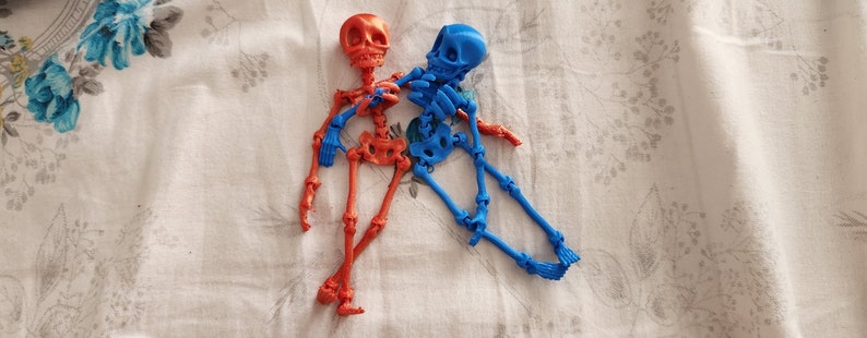 May include: Two articulated skeleton figures, one red and one blue, are lying on a white fabric surface with a floral pattern. The skeletons are posed in a relaxed, almost playful manner.