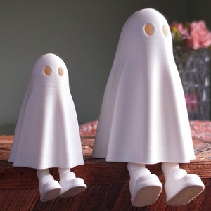 May include: Two white ghost figurines with round eyes and simple legs, sitting on a wooden surface.