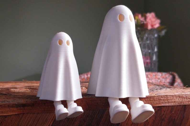 May include: Two white ghost figurines with round eyes and simple legs, sitting on a wooden surface.