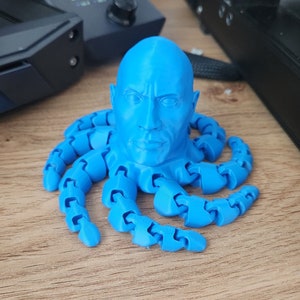 May include: A blue 3D printed sculpture of a person's head with a muscular build, resembling Dwayne 'The Rock' Johnson, with eight blue articulated octopus tentacles.