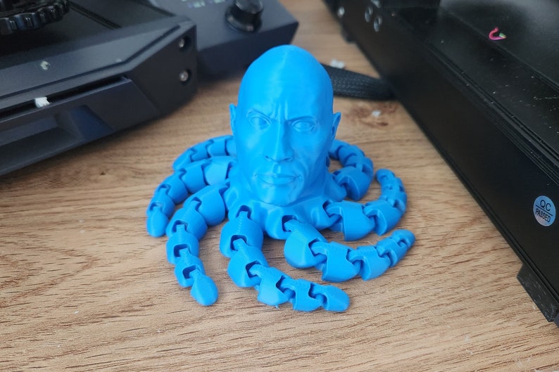 May include: A blue 3D printed sculpture of a person's head with a muscular build, resembling Dwayne 'The Rock' Johnson, with eight blue articulated octopus tentacles.
