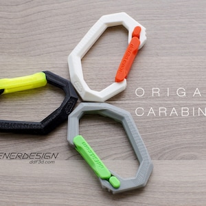 May include: Three origami carabiners in different colors: black with a yellow latch, white with an orange latch, and gray with a green latch. The carabiners are arranged in a circle on a wooden surface. The text "ORIGAMI CARABINER" is printed in black on the wooden surface.