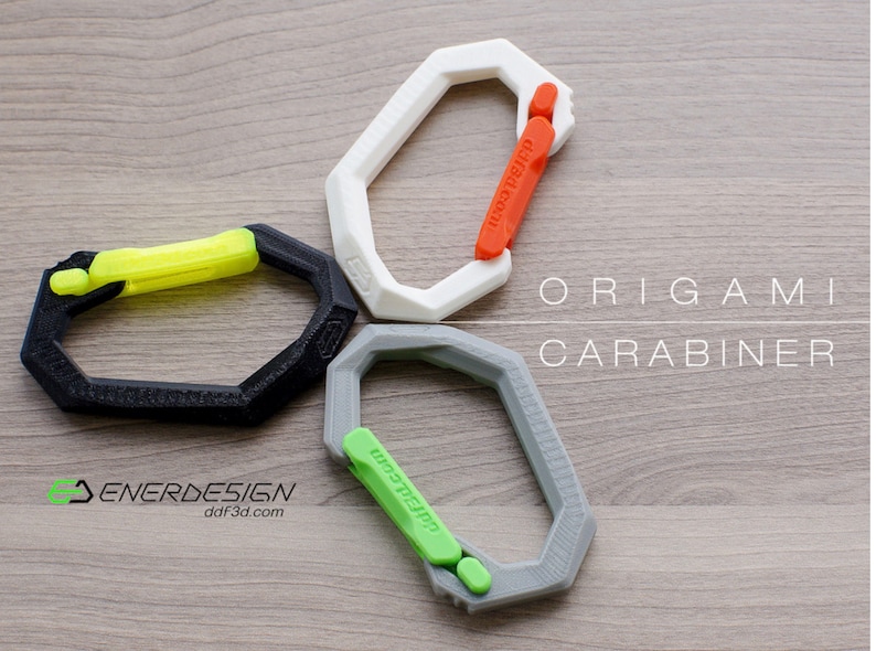 May include: Three origami carabiners in different colors: black with a yellow latch, white with an orange latch, and gray with a green latch. The carabiners are arranged in a circle on a wooden surface. The text "ORIGAMI CARABINER" is printed in black on the wooden surface.