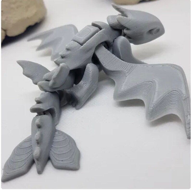 May include: A gray 3D printed dragon figurine with articulated wings and tail. The dragon has a detailed, textured surface.