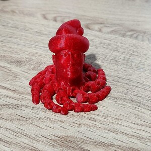 Puede incluir: Una figura roja impresa en 3D de una persona con un gorro de Pap&aacute; Noel y una larga barba. La figura est&aacute; de pie sobre una superficie de madera.