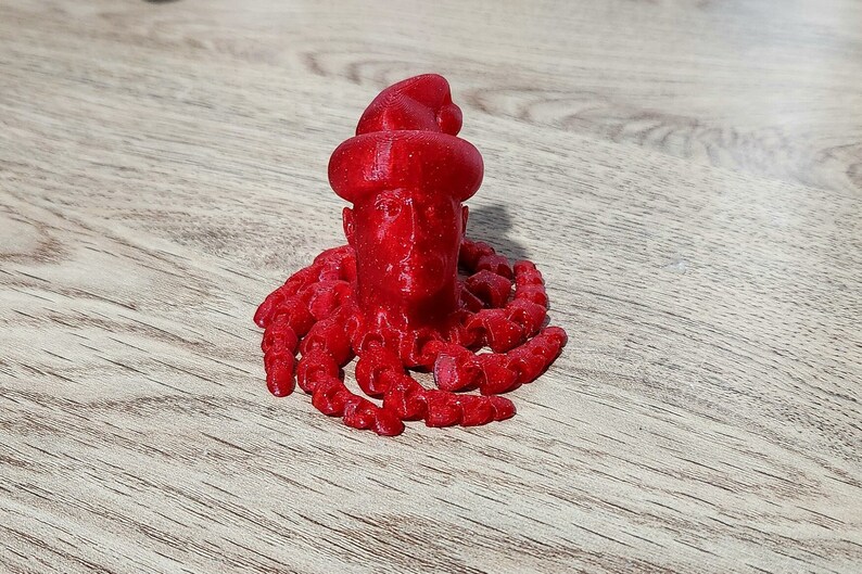 Puede incluir: Una figura roja impresa en 3D de una persona con un gorro de Pap&aacute; Noel y una larga barba. La figura est&aacute; de pie sobre una superficie de madera.