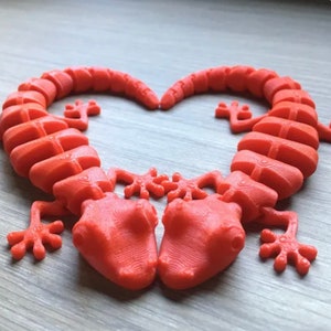 Może przedstawiać: Koralowa, drukowana w 3D rzeźba w kształcie serca, złożona z dw&oacute;ch stylizowanych figurek gekona. Każdy gekon ma szczeg&oacute;łowe, teksturowane ciało i cztery przegubowe nogi. Rzeźba jest przeznaczona jako przedmiot dekoracyjny.