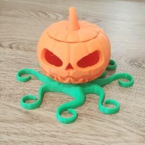 May include: A 3D printed orange pumpkin with a carved face sits on a green octopus-shaped base. The pumpkin has a small stem on top and is hollowed out.