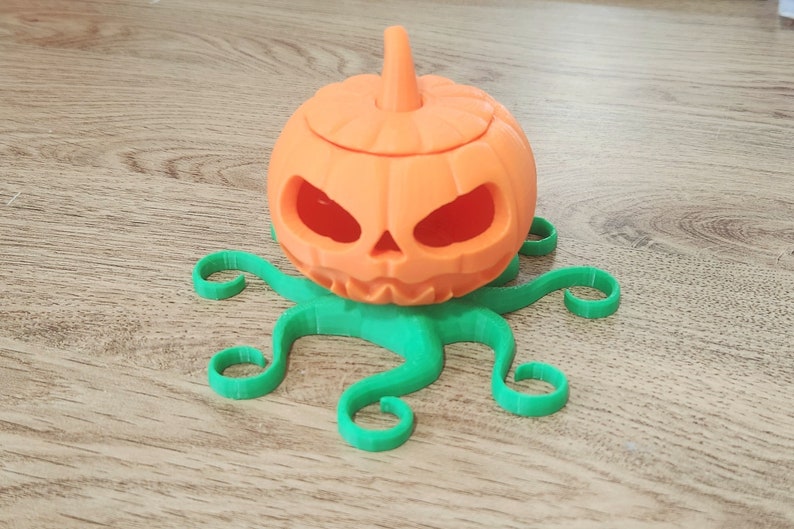 May include: A 3D printed orange pumpkin with a carved face sits on a green octopus-shaped base. The pumpkin has a small stem on top and is hollowed out.