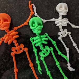 May include: Three articulated skeleton figurines, one orange, one green, and one white. The skeletons are posed in a playful, dancing manner.