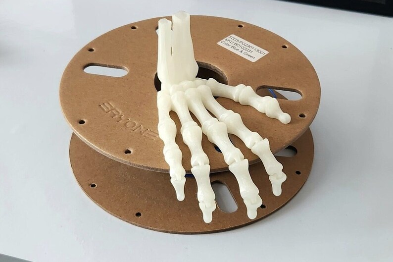 May include: A white plastic 3D printed model of a human hand skeleton on a brown spool. The spool has the text "ERYONE" printed on it. The label on the spool reads "PLA1.75 200G 1.0017 SKU: 8011522010001 Color: Blue & Green"