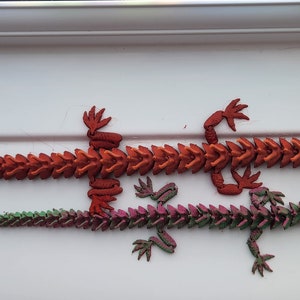 May include: Two 3D printed articulated dragon figurines. The larger dragon is red and the smaller dragon is green and purple.