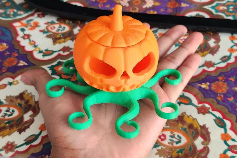 May include: A 3D printed orange pumpkin with a green octopus base. The pumpkin has a carved face and a stem on top.
