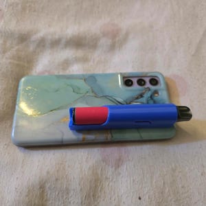 K&ouml;nnte beinhalten: Ein blauer und roter Vape Pen liegt auf einer Smartphone-H&uuml;lle. Die Handyh&uuml;lle hat ein Marmormuster in Blau-, Gr&uuml;n- und Goldt&ouml;nen. Der Vape Pen ist blau mit einer roten Kartusche.