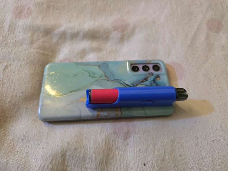 K&ouml;nnte beinhalten: Ein blauer und roter Vape Pen liegt auf einer Smartphone-H&uuml;lle. Die Handyh&uuml;lle hat ein Marmormuster in Blau-, Gr&uuml;n- und Goldt&ouml;nen. Der Vape Pen ist blau mit einer roten Kartusche.