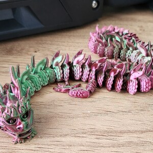 May include: A green and pink 3D printed dragon figurine with a detailed head and scales. The dragon is curled up in a coil shape.