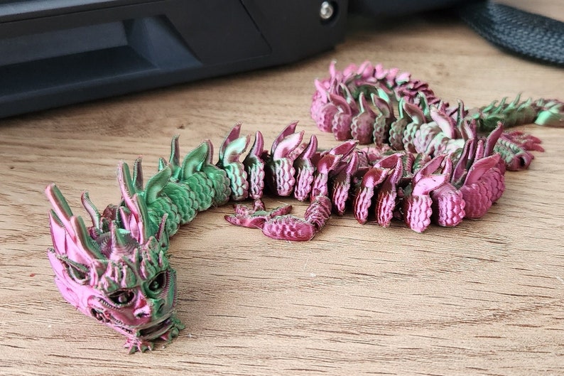 May include: A green and pink 3D printed dragon figurine with a detailed head and scales. The dragon is curled up in a coil shape.