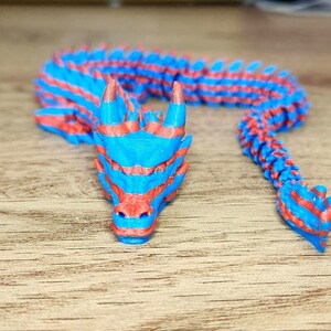 Puede incluir: Una figurita de drag&oacute;n impresa en 3D con rayas azules y rojas. El drag&oacute;n est&aacute; enroscado con la cabeza apoyada en el suelo.
