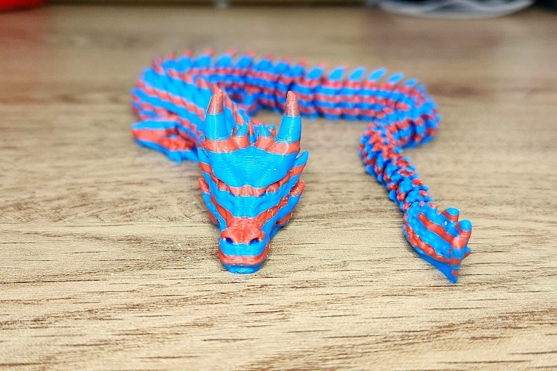 Puede incluir: Una figurita de drag&oacute;n impresa en 3D con rayas azules y rojas. El drag&oacute;n est&aacute; enroscado con la cabeza apoyada en el suelo.