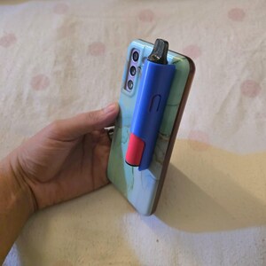 K&ouml;nnte beinhalten: Ein blau-roter Vape-Pen an einem Smartphone befestigt. Das Telefon hat eine hellblaue H&uuml;lle mit Marmormuster. Der Vape-Pen ist blau mit rotem Akzent und schwarzem Mundst&uuml;ck. Das Telefon wird in einer Hand gehalten.