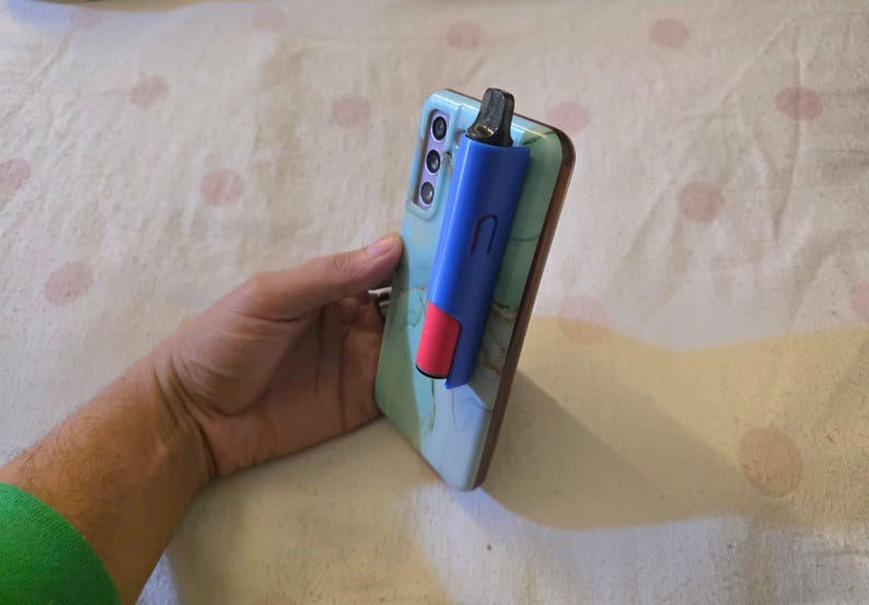 K&ouml;nnte beinhalten: Ein blau-roter Vape-Pen an einem Smartphone befestigt. Das Telefon hat eine hellblaue H&uuml;lle mit Marmormuster. Der Vape-Pen ist blau mit rotem Akzent und schwarzem Mundst&uuml;ck. Das Telefon wird in einer Hand gehalten.