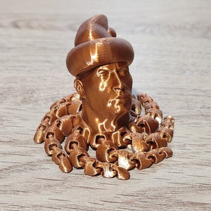 Puede incluir: Un busto impreso en 3D de una persona con un sombrero, con una cadena de peque&ntilde;os anillos unidos alrededor del cuello. El busto y la cadena son de color cobre met&aacute;lico.
