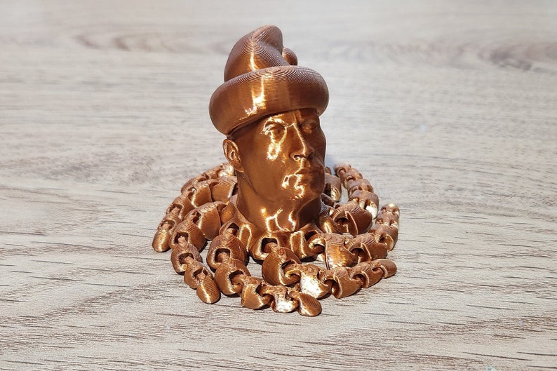 Puede incluir: Un busto impreso en 3D de una persona con un sombrero, con una cadena de peque&ntilde;os anillos unidos alrededor del cuello. El busto y la cadena son de color cobre met&aacute;lico.