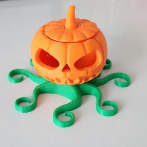 May include: A 3D printed orange pumpkin with a carved face sits on a green, 3D printed octopus-like base.