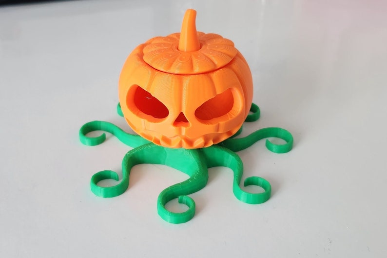 May include: A 3D printed orange pumpkin with a carved face sits on a green, 3D printed octopus-like base.