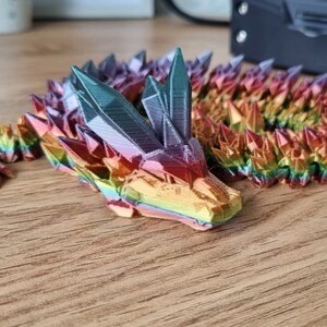 May include: A rainbow-colored 3D printed dragon figurine with a crystal-like texture. The dragon is curled up with its head resting on the ground.