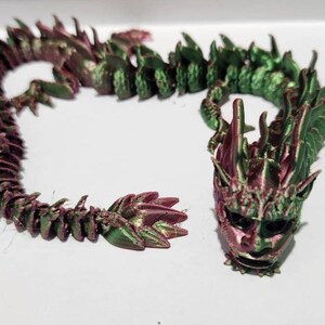 May include: A 3D printed dragon figurine with a green and pink iridescent finish. The dragon is curled up in a coil shape with its head facing forward. The dragon's head has a detailed face with sharp teeth and horns.