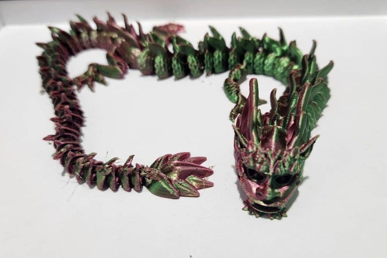 May include: A 3D printed dragon figurine with a green and pink iridescent finish. The dragon is curled up in a coil shape with its head facing forward. The dragon's head has a detailed face with sharp teeth and horns.