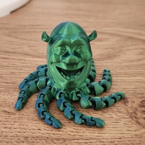 Puede incluir: Una figura verde impresa en 3D de Shrek, el personaje de la pel&iacute;cula Shrek, con una cara sonriente y ocho tent&aacute;culos.