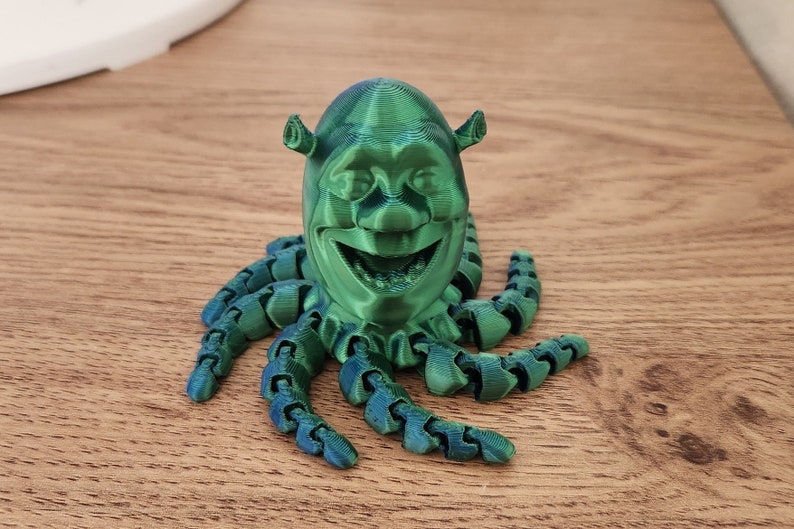 Puede incluir: Una figura verde impresa en 3D de Shrek, el personaje de la pel&iacute;cula Shrek, con una cara sonriente y ocho tent&aacute;culos.