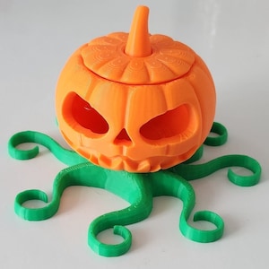 May include: A 3D printed orange pumpkin with a carved face and a green stem, sitting on a green, 3D printed base with six tentacles.