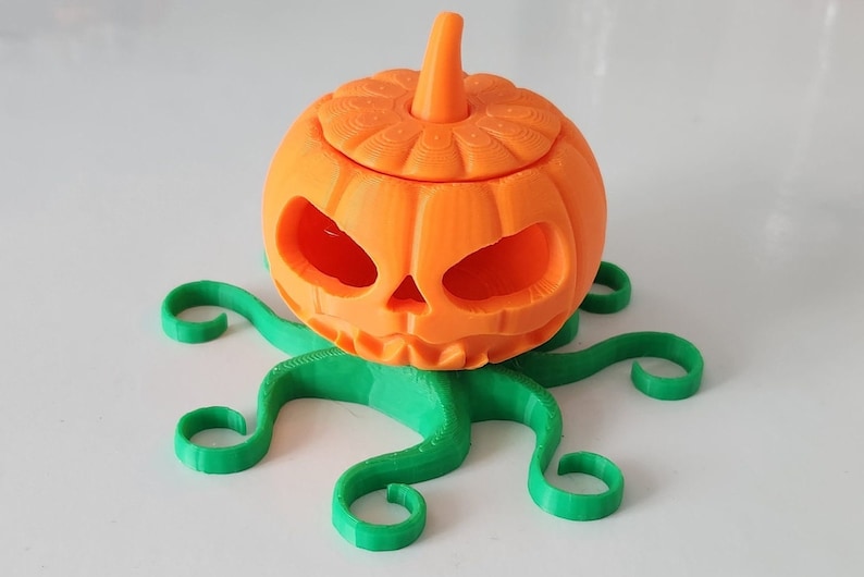 May include: A 3D printed orange pumpkin with a carved face and a green stem, sitting on a green, 3D printed base with six tentacles.